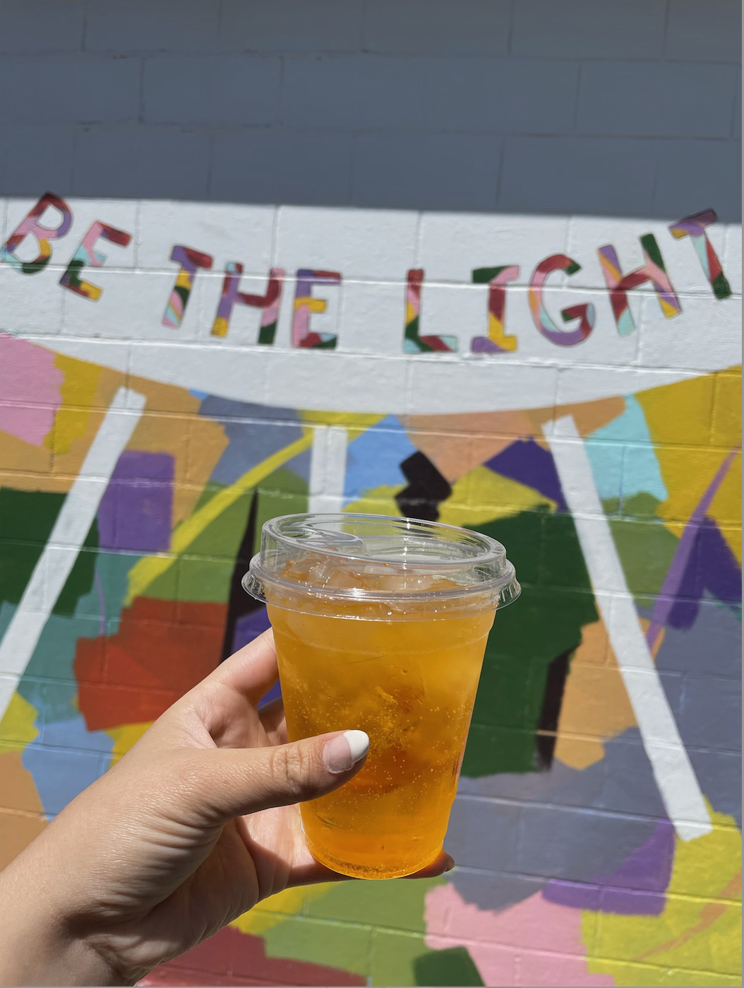 An iced sparkling drink held up to a colorful Be the Light mural