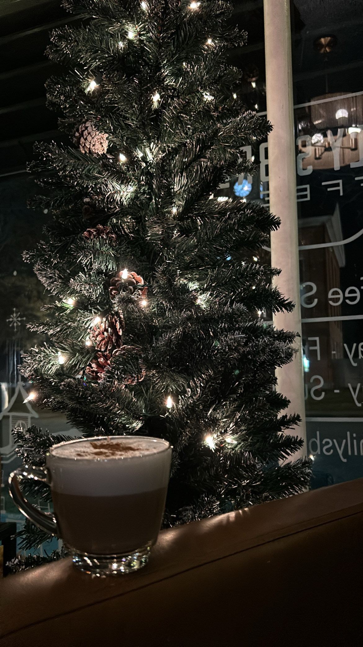 A latte in a glass cup beside a lit Christmas tree in the shop window